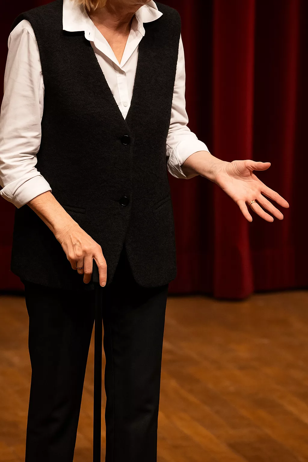 Silhouette d’une comédienne sur scène, cadrée du cou aux jambes, devant un rideau rouge. Ses mains expressives accompagnent un geste, tandis qu’elle tient un micro. Lumière chaude et ambiance de théâtre parisien. Aucun visage visible.